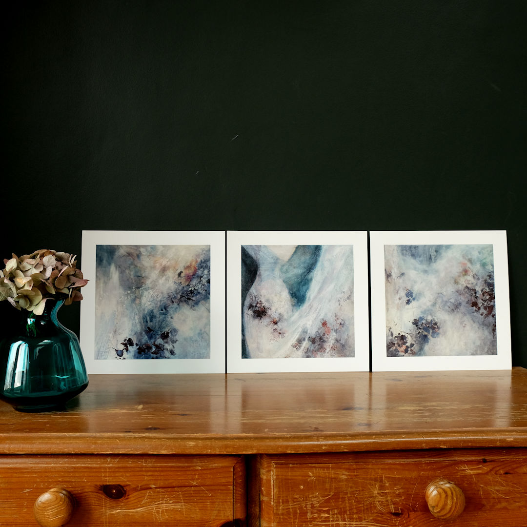 Ghost of Winter 1, 2 & 3 giclee prints shown stood against a dark wall on a desk with a glass vase to the side with a faded hydrangea flower